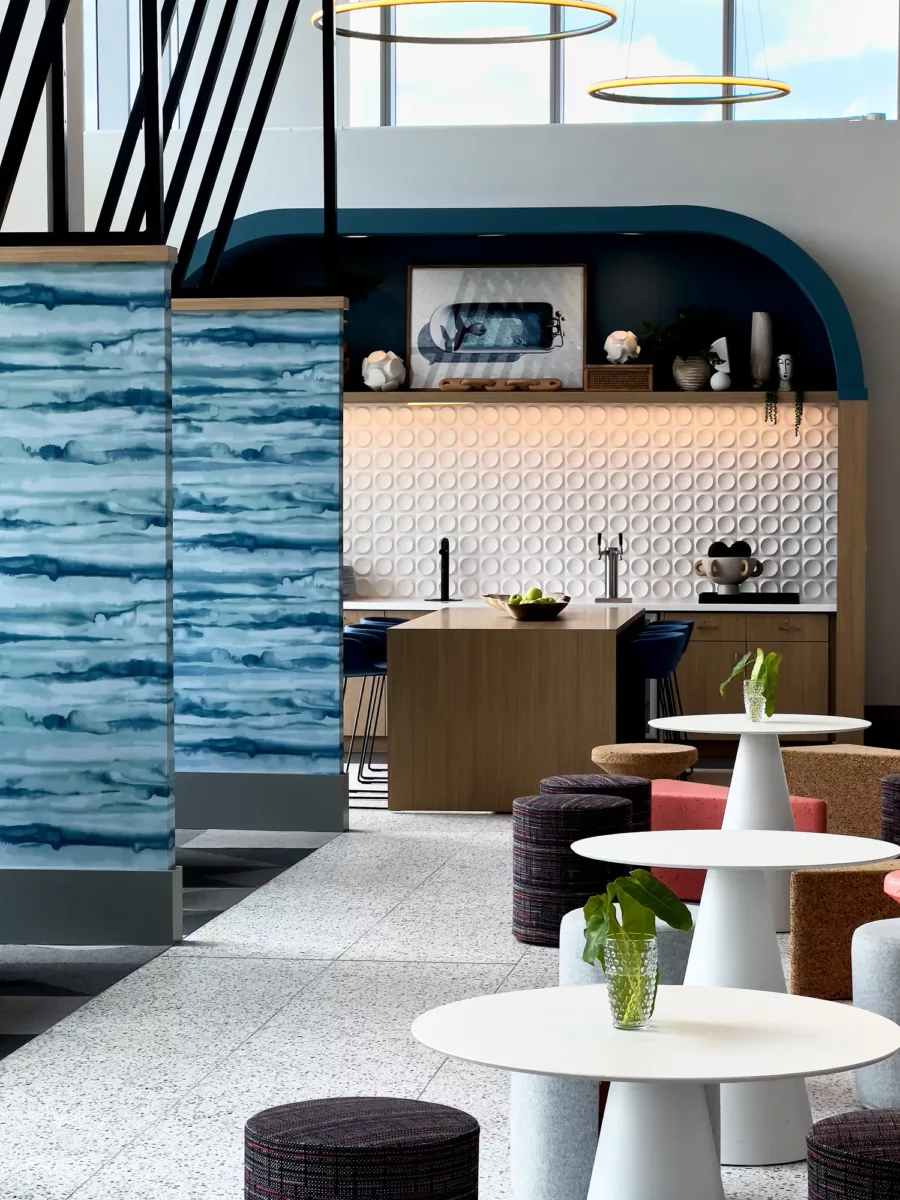 Community lounge with round tables, upholstered stools, and modern kitchenette backdrop.