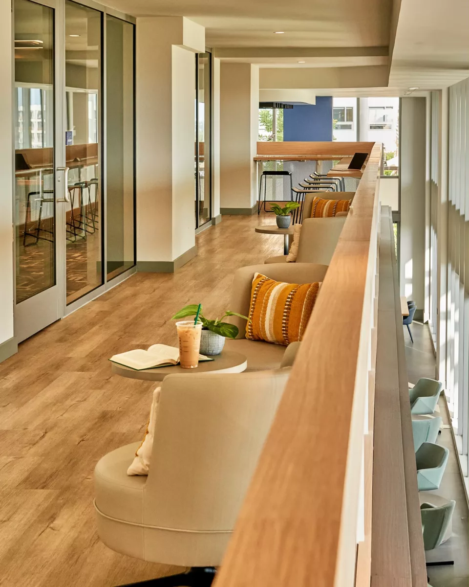 Upper‑level corridor with lounge seating, wood floors, glass offices, and natural light.