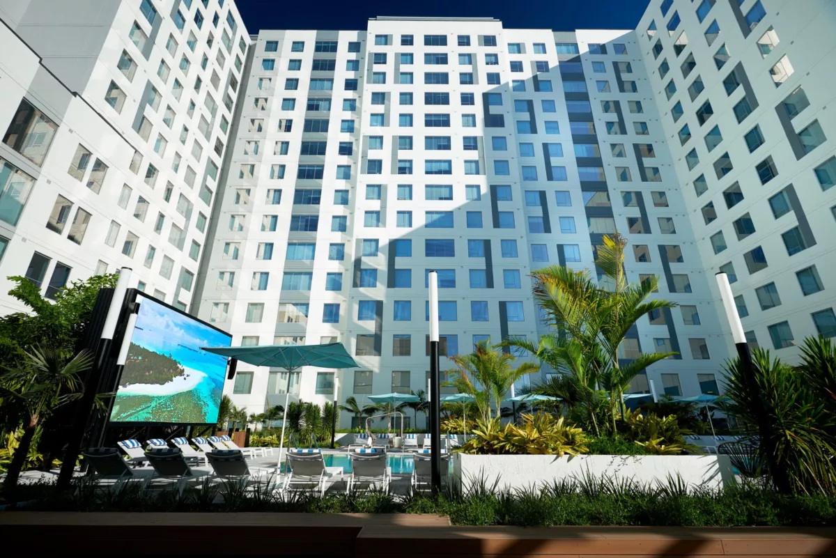 Outdoor pool courtyard with lounge chairs, umbrellas, and large screen