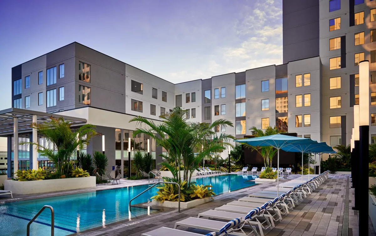 Resort-style pool area at dusk with lounge chairs, umbrellas, and illuminated buildings