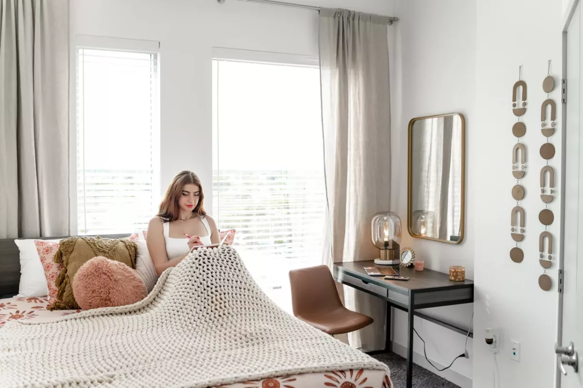 Bedroom interior with bed, textured bedding, writing desk, mirror, and large windows.