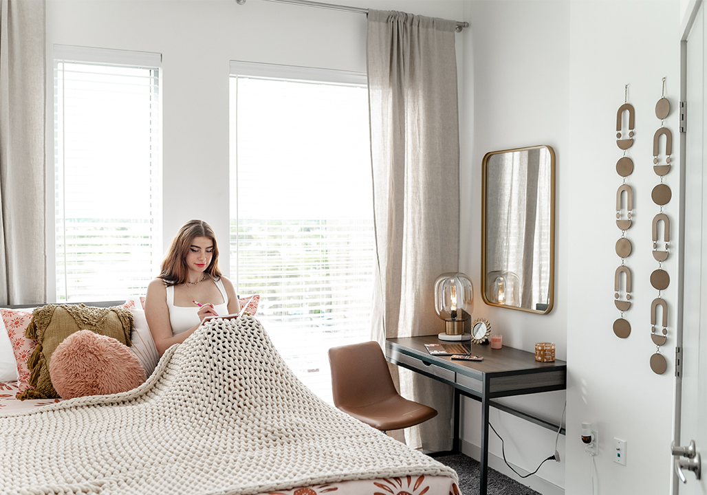 A woman sits on a bed with a book in a bedroom with large windows and white walls