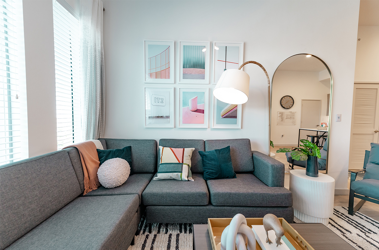 Living room with large windows and white walls with colorful framed photographs, a gray couch with pillows, and a mirror in the background