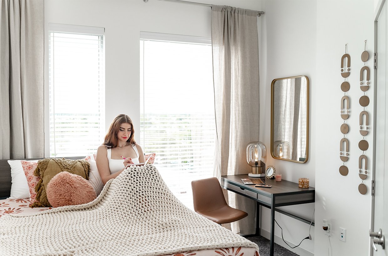 A woman sits on a bed with a book in a bedroom with large windows and white walls